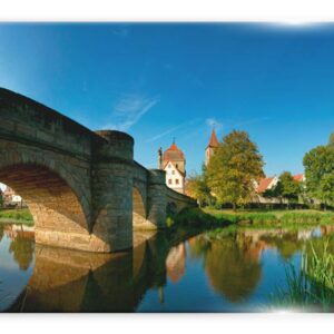 תמונה של גשר אבן עתיק מעל נהר, עם נוף של ע treesים ירוקים ובתים בסגנון כפרי ברקע תחת שמיים בהירים.